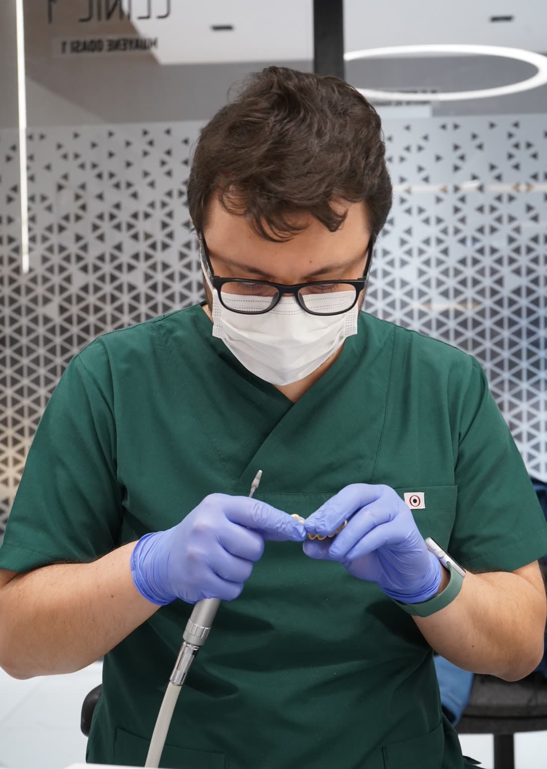 Dentista no consultório, de jaleco verde, preparando uma prótese